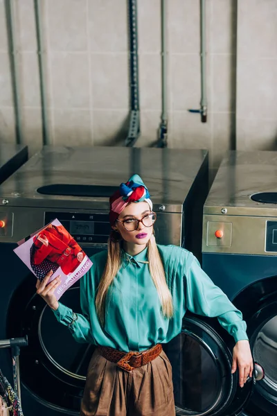 Mujer joven y de moda en turbante y gafas celebración revista en lavandería - foto de stock