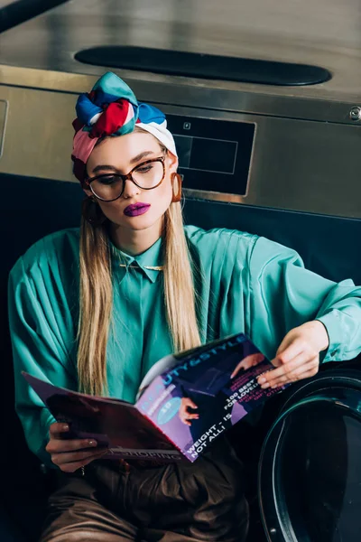 Élégant jeune femme en turban et lunettes de lecture magazine en laverie automatique — Photo de stock