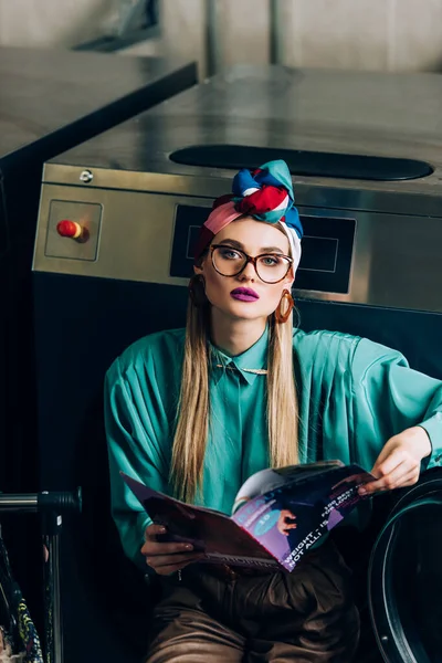 Élégant jeune femme en turban et lunettes tenant magazine en laverie automatique — Photo de stock