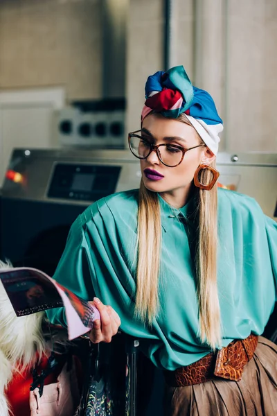 Jeune et élégante femme dans des lunettes et turban lecture magazine en laverie automatique — Photo de stock