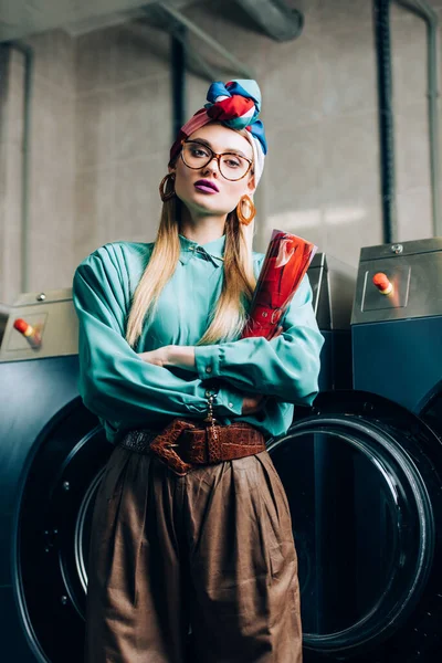 Trendfrau mit Brille und Turban steht mit verschränkten Armen im öffentlichen Waschsalon und hält Magazin in der Hand — Stockfoto