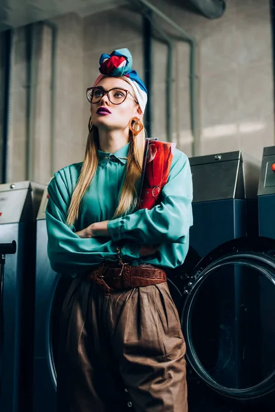 Joven mujer de moda en gafas y turbante de pie con los brazos cruzados y la celebración de revista en la lavandería pública - foto de stock