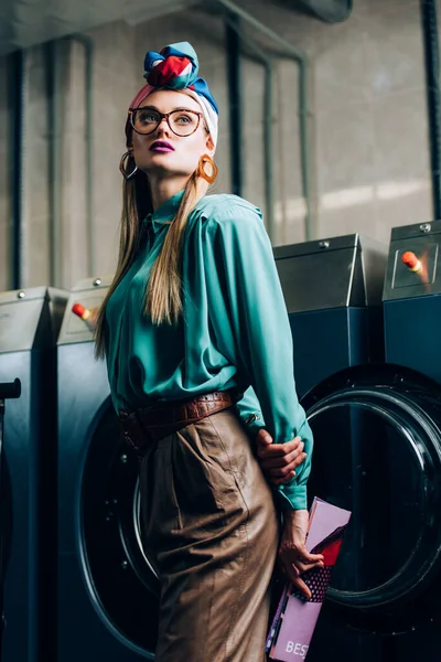 Femme à la mode dans les lunettes et turban tenant magazine dans la laverie automatique publique — Photo de stock