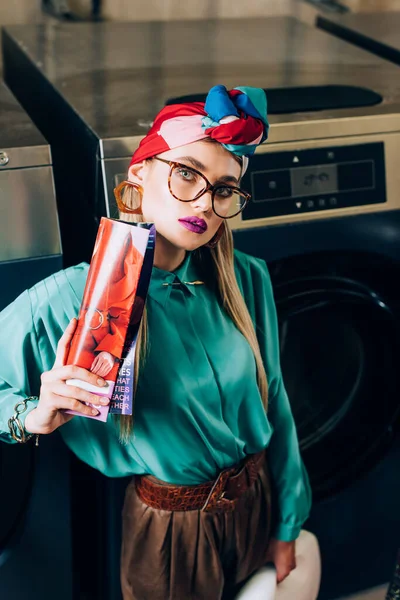 Young woman in glasses and turban holding magazine in public laundromat — Stock Photo