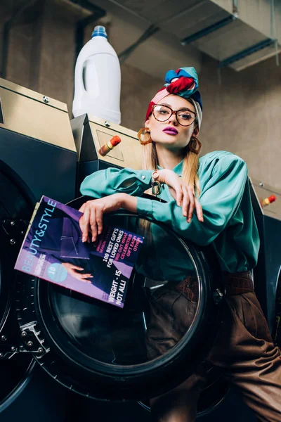 Low angle view of young woman in glasses and turban holding magazine near washing machines in public laundromat — Stock Photo