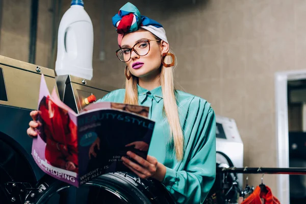 Woman in glasses and turban holding magazine in public laundromat — Stock Photo