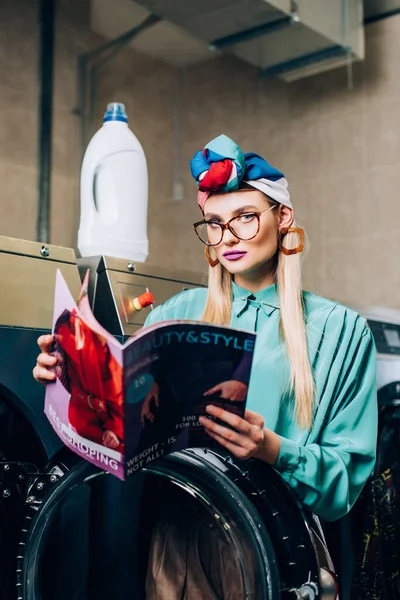 Woman in glasses and turban holding magazine in laundromat — Stock Photo