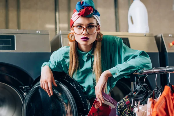 Woman in glasses and turban holding magazine near modern washing machine in public laundromat — Stock Photo