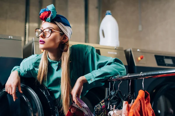 Femme à la mode dans les lunettes et turban tenant magazine près de la machine à laver moderne — Photo de stock