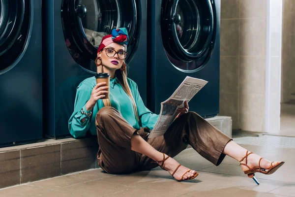 Trendy woman in glasses and turban holding paper cup and newspaper while sitting in laundromat — Stock Photo