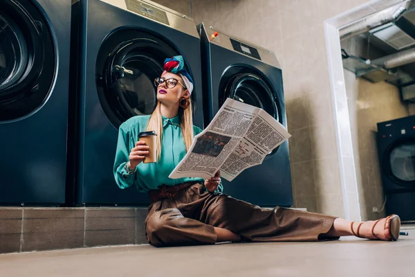 Donna alla moda in bicchieri e turbante in possesso di tazza di carta e giornale mentre seduto sul pavimento in lavanderia — Foto stock