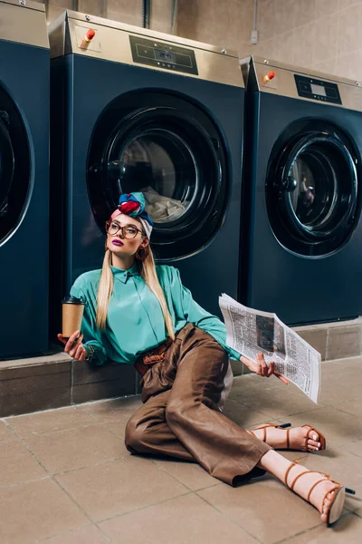 Junge Frau mit Brille und Turban hält Pappbecher und Zeitung in der Hand, während sie im Waschsalon auf dem Boden sitzt — Stockfoto
