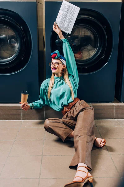 Glückliche Frau mit Brille und Turban, die Pappbecher und Zeitung in der Hand hält, während sie im Waschsalon auf dem Boden sitzt — Stockfoto