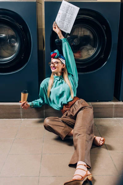 Mujer feliz en gafas y turbante sosteniendo taza de papel y periódico mientras se sienta en el suelo en la lavandería - foto de stock