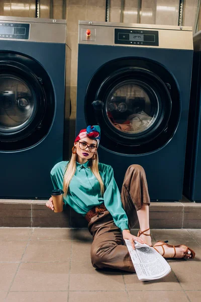 Junge Frau mit Brille und Turban hält Pappbecher und Zeitung in der Hand, während sie im Waschsalon auf dem Boden sitzt — Stockfoto
