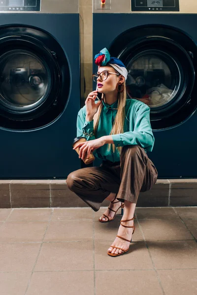 Jeune femme en lunettes et turban parler sur smartphone et tenant tasse de papier dans la laverie automatique — Photo de stock