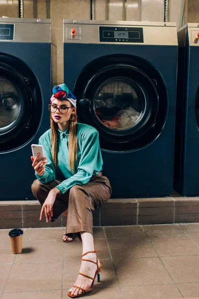 Jeune femme en lunettes et turban tenant smartphone près de tasse de papier dans la laverie automatique — Photo de stock