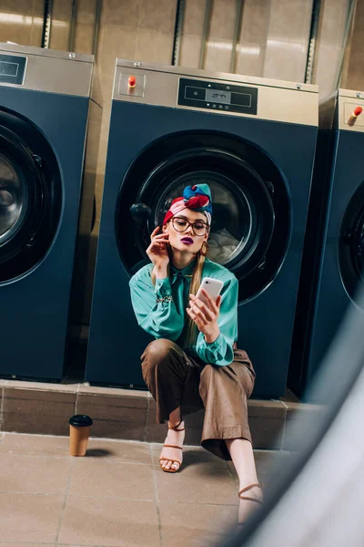 Mujer joven en gafas y turbante mirando teléfono inteligente cerca de taza de papel en lavandería - foto de stock