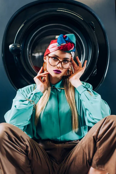 Jeune femme en lunettes et turban regardant la caméra près de la machine à laver — Photo de stock