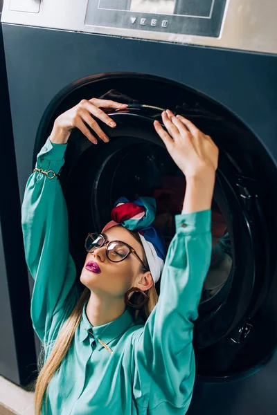 Stylish young woman in glasses and turban touching door of washing machine in laundromat — Stock Photo