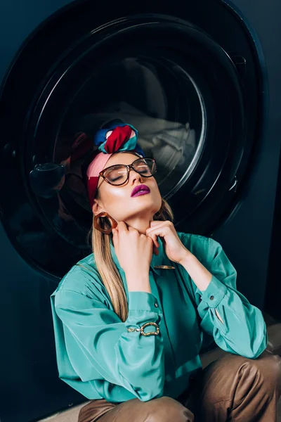 Стильная молодая женщина в очках и тюрбан сидит с закрытыми глазами возле стиральной машины — стоковое фото
