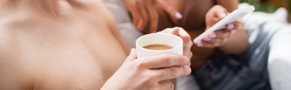 Teilbild eines Mannes mit Kaffeetasse in der Nähe seiner Freundin, der mit dem Finger auf das Smartphone zeigt, auf verschwommenem Hintergrund, Banner — Stockfoto