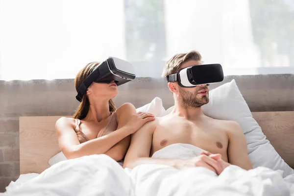 Mujer joven tocando novio sin camisa mientras usa auriculares vr en la cama - foto de stock