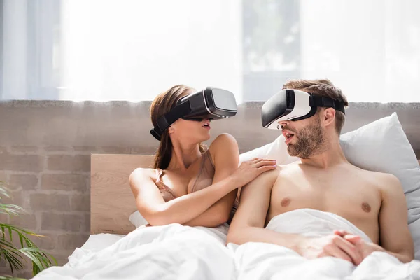 Couple excité se regardant tout en étant couché dans le lit et en utilisant des casques vr — Photo de stock