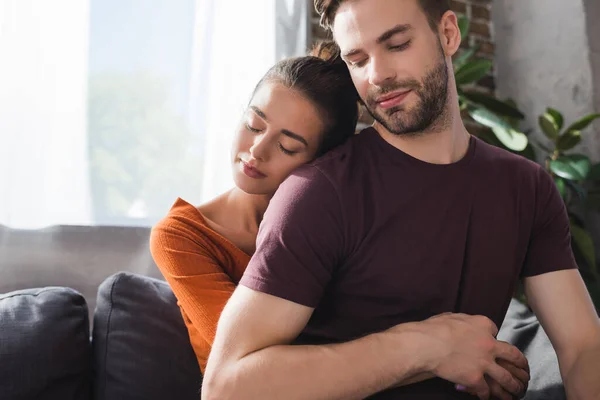 Mujer tierna con los ojos cerrados abrazando al hombre amado desde la espalda mientras está sentado en el sofá - foto de stock