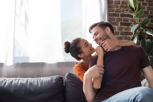 Cheerful woman touching face of happy boyfriend while sitting on sofa at home — Stock Photo