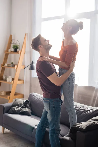 Side view of young man hugging girlfriend standing on sofa at home — Stock Photo