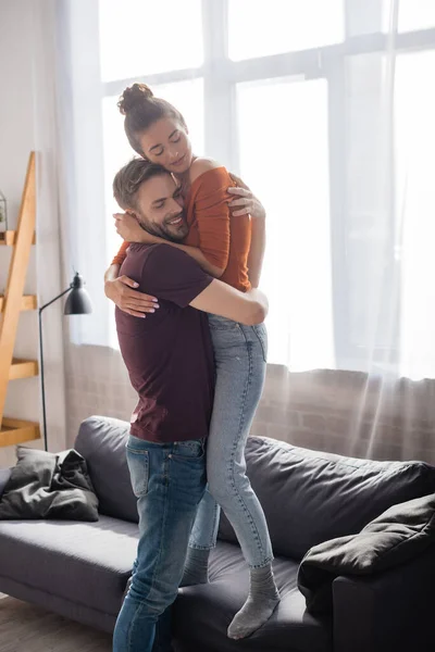 Happy man hugging beloved girlfriend standing on sofa at home — Stock Photo