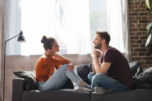 Vista lateral do jovem casal sentado no sofá e falando enquanto olha um para o outro — Fotografia de Stock