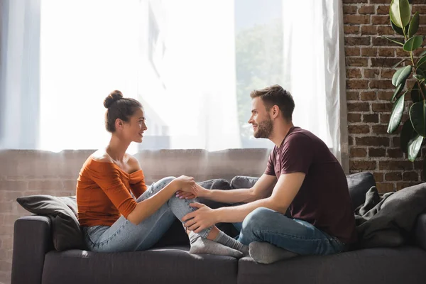 Vista lateral de la joven pareja hablando mientras está sentada en el sofá y cogida de la mano - foto de stock