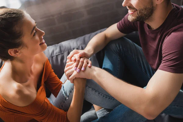 Sonriente pareja cogida de la mano y hablando mientras se sienta en el sofá - foto de stock