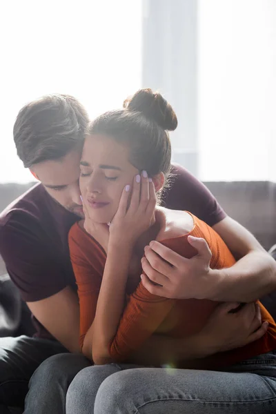 Zarter Mann umarmt weinende Freundin, die Gesicht berührt, während sie auf Sofa sitzt — Stockfoto