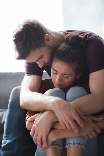 Joven abrazando y calmando a la amada mujer sentada en el sofá con los ojos cerrados - foto de stock