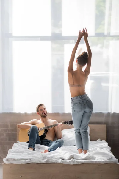 Вид ззаду молодої жінки в джинсах і бюстгальтер танцює на ліжку біля хлопця, граючи на гітарі — стокове фото