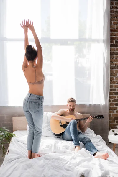 Rückansicht der verführerischen Frau, die mit erhobenen Händen neben ihrem Freund tanzt und im Bett Gitarre spielt — Stockfoto