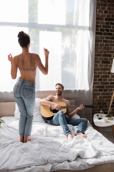 Back view de mulher sexy em jeans e sutiã dançando na cama perto de homem alegre tocando guitarra — Fotografia de Stock