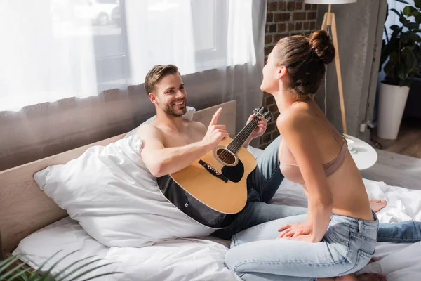 Sonriente hombre sosteniendo la guitarra y señalando con el dedo cerca de la novia en el sujetador sentado en la cama - foto de stock