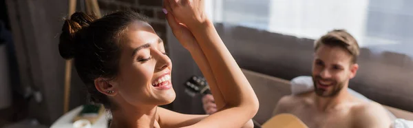 Excitada mujer bailando con los ojos cerrados cerca de novio tocando la guitarra sobre fondo borroso, pancarta - foto de stock