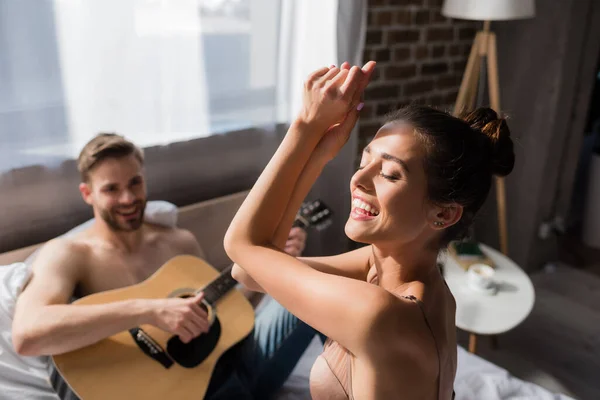 Eccitata donna in reggiseno danza con gli occhi chiusi vicino fidanzato suonare la chitarra su sfondo sfocato — Foto stock