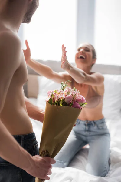 Homem sem camisa segurando flores perto de namorada animado com as mãos estendidas no fundo borrado — Fotografia de Stock