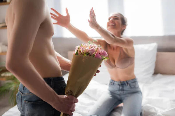 Hombre sin camisa sosteniendo flores cerca de la mujer excitada con las manos extendidas sobre fondo borroso - foto de stock