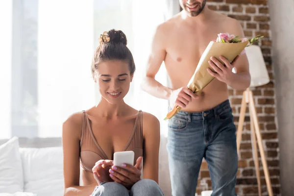 Lächelnde, sexy Frau plaudert auf Smartphone neben hemdlosem Mann mit Blumenstrauß auf verschwommenem Hintergrund — Stockfoto