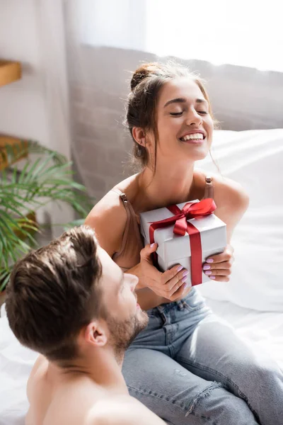 Femme excitée avec les yeux fermés tenant boîte cadeau près de l'homme sur le premier plan flou — Photo de stock