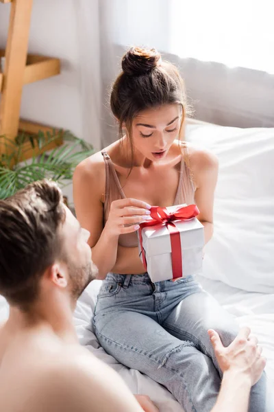 Здивована дівчина в джинсах і бюстгальтер відкриває подарункову коробку біля чоловіка на розмитому передньому плані — стокове фото