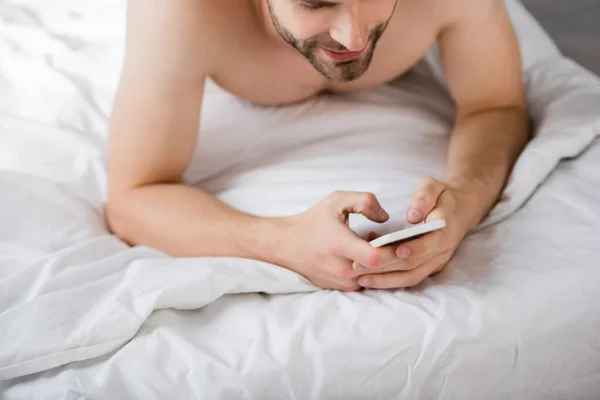 Обрезанный вид молодого человека, лежащего в постели и болтающего на смартфоне — стоковое фото