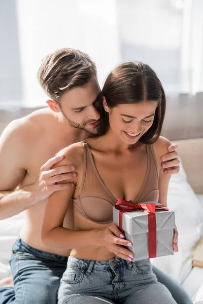 Shirtless man hugging sexy girlfriend holding gift box in bedroom — Stock Photo